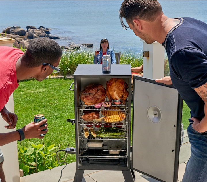 Electric smoker at cookout with 2 people looking at food inside
