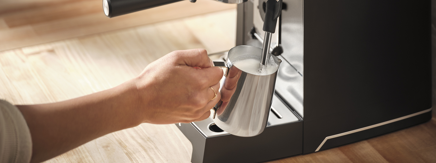 Hand holding milk carafe to frothing wand.